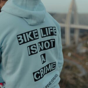 Man in gray hoodie with message 'Bike Life Is Not A Crime' outdoors near a bridge.
